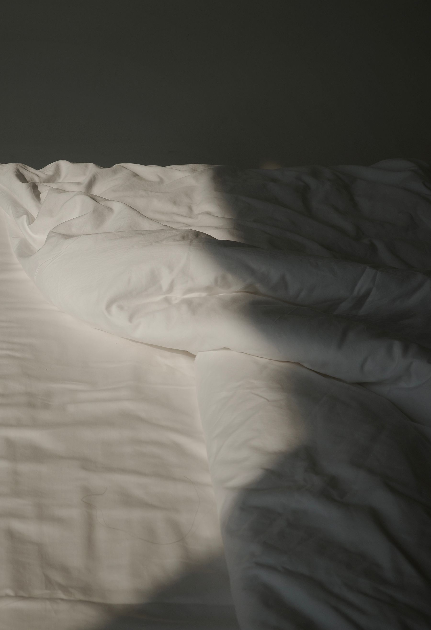 White bed linen in low side light, folds and creases across a duvet and sheet