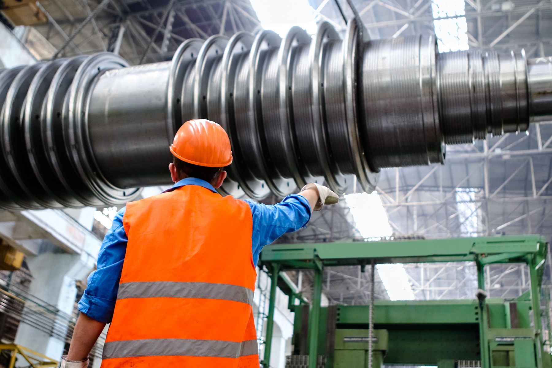 Engineer in a hard hat and safety vest inspecting a large machined steel shaft inside an industrial plant, seen from behind