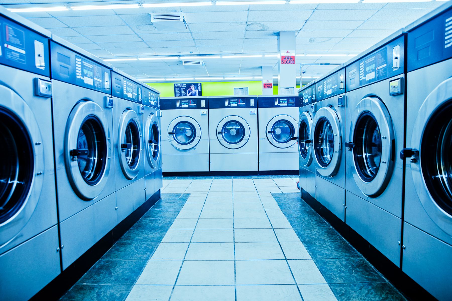 Rows of front-load commercial washing machines facing each other across a tiled laundry aisle