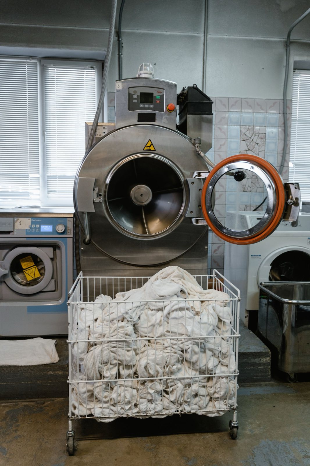 Industrial washer-extractor with its door open beside a wire trolley filled with linen, in a working laundry room