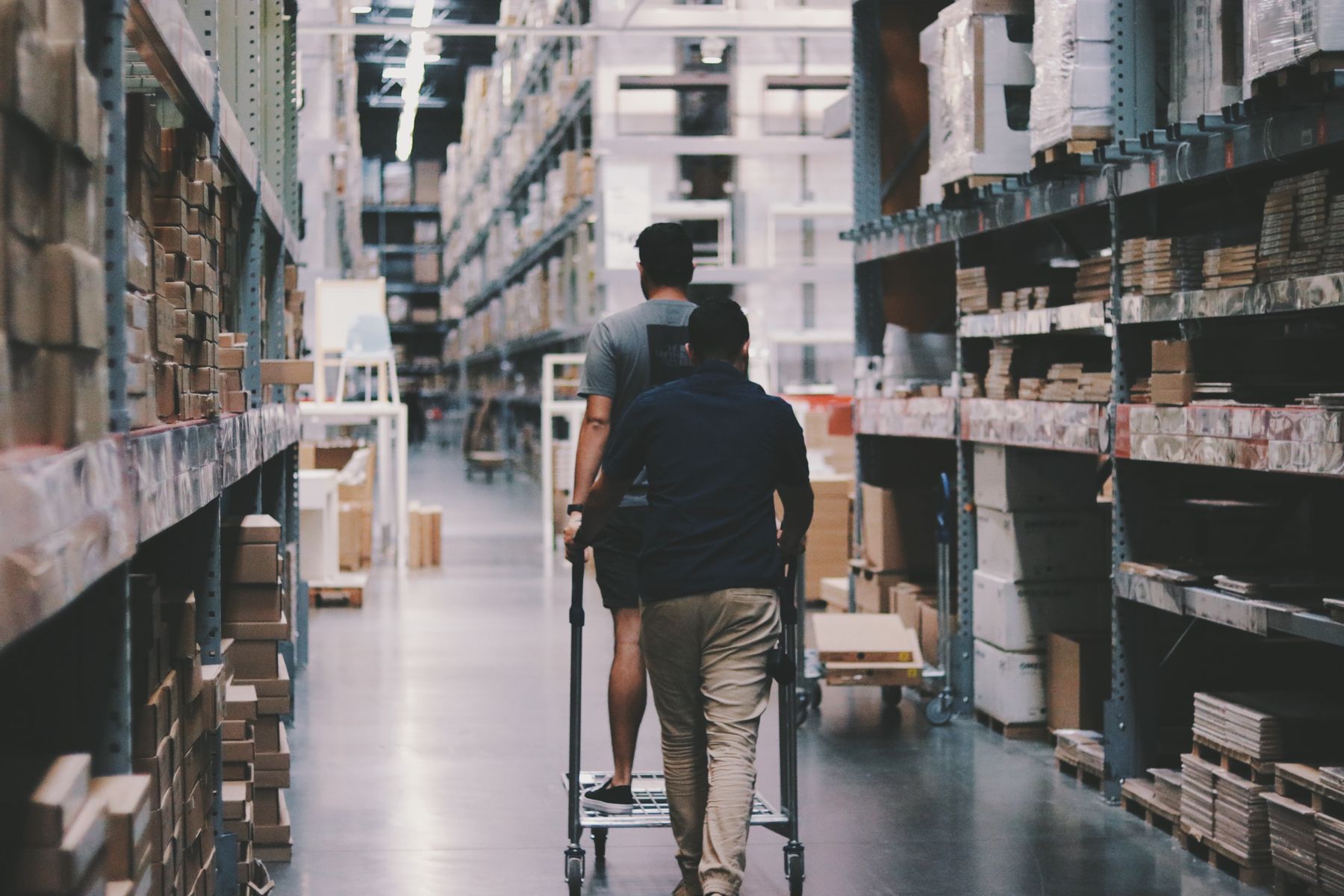 Two warehouse workers pushing a flatbed cart down a racked aisle, seen from behind